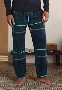 Mies, jolla on yllään tummanvihreät ja tummansiniset ruudulliset pyjamahousut, seisoo paljain jaloin kuvioidulla matolla kodikkaassa huoneessa.