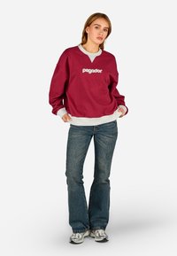 Burgundi színű kapucnis pulóver fehér logóval, szürke gallérral, hosszú ujjakkal, strukturált anyagból, kék farmernadrággal és szürke sportcipővel párosítva.