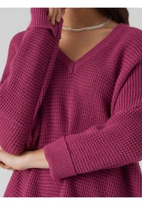 Femme portant un pull en tricot gaufré magenta à col en V et manches longues, tenant une main près de son visage, portant un collier chaîne argenté.