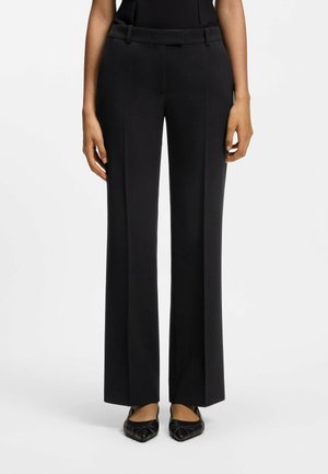 Kobieta w czarnych szerokich spodniach garniturowych i czarnych balerinach, stojąca na tle jednolitego białego tła.