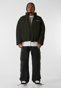 Sort pufferjakke med høj krave og lynlås, lagd over en grå sweater og en hvid T-shirt, kombineret med sorte cargo-bukser og sneakers.