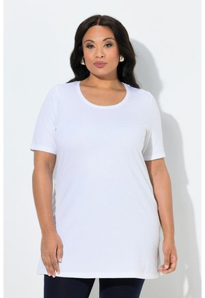 Donna con capelli lunghi e scuri che indossa orecchini a cerchio dorati e una maglietta bianca a maniche corte semplice, in piedi davanti a uno sfondo bianco.