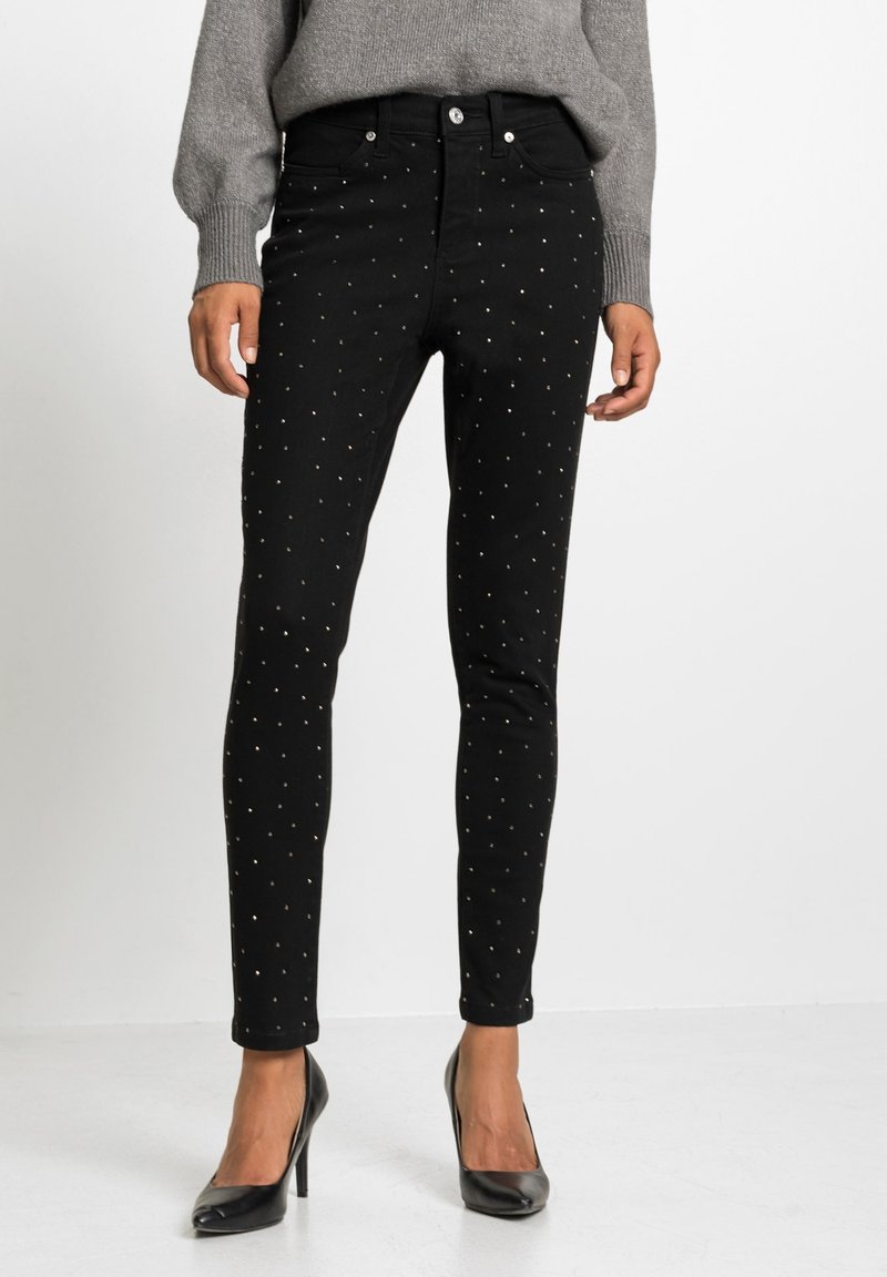 Pantalon noir ajusté avec de petits clous argentés, porté avec un pull gris et des chaussures à talons noirs, debout sur un sol blanc.