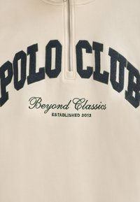 Felpa crema con zip a un quarto, testo ricamato scuro che legge "POLO CLUB" e "Beyond Classics" in verde, con l'anno di fondazione 2012.