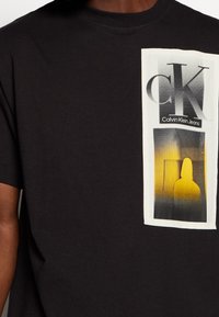 Schwarzes Baumwoll-T-Shirt mit einem grafischen Druck in Weiß und Gelb, das "CK" und "Calvin Klein Jeans" mit einem Silhouetten-Design zeigt.