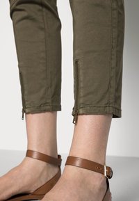 Olive green cropped pants made of cotton, featuring brass zipper details at the cuffs. Worn with brown strappy sandals.