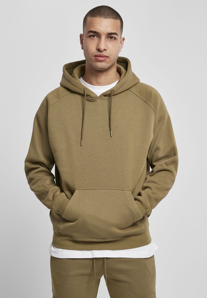 Man standing with hands in pocket of olive green hoodie, wearing matching pants and a white shirt underneath, neutral background.