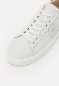 Adidași albi cu șireturi, cu un superior din piele netedă, vârf rotunjit și talpă din cauciuc texturat; prezintă un mic logo embosat pe lateral.