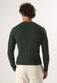 Ciemnozielony sweter w splot kablowy z okrągłym dekoltem i ściągaczami, z widocznymi teksturowanymi paskami. Model ma na sobie jasne spodnie.