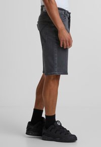 Shorts en denim noir avec une coupe droite, comportant une étiquette à la taille, cinq poches et des détails de couture. Associés à des baskets noires et des chaussettes.
