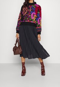 Gebreide trui met kleurrijke bloemenpatronen, gecombineerd met een zwarte geplooide rok. Accessoires zijn een bruine tas en glanzende bruine enkellaarsjes.