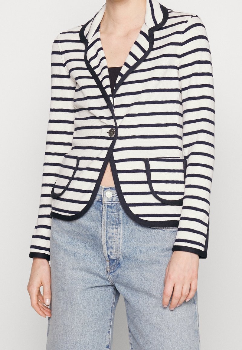 Personne portant un blazer rayé blanc et marine avec une bordure noire, un bouton fermé, associé à un jean taille haute bleu clair.