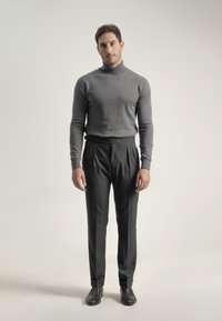 Hombre de pie con un suéter gris de cuello alto, pantalones gris oscuro y zapatos negros contra un fondo claro y liso.