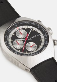 Montre Timex avec un boîtier en acier inoxydable, un cadran noir, trois sous-cadrans, des accents rouges, une échelle tachymétrique et un bracelet en caoutchouc noir texturé.