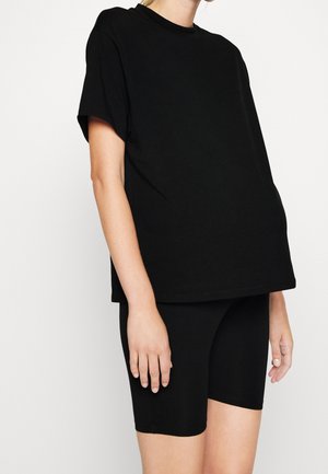 Osoba ubrana w luźną czarną koszulkę z krótkim rękawem w połączeniu z dopasowanymi czarnymi spodenek rowerowych do kolan na jednolitym tle.