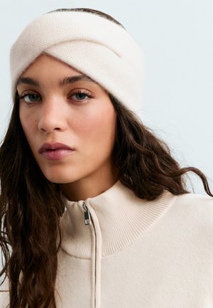Une femme aux longs cheveux foncés porte un bandeau en maille couleur crème et un pull à col zippé sur un fond clair.
