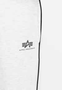 Svetlosive trenirke imajo črno črto ob strani in natisnjen logotip "ALPHA INDUSTRIES" blizu žepa. Mehka tekstura.