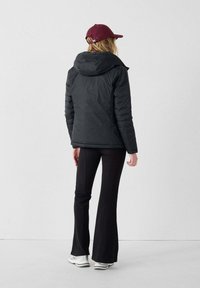 Zwarte pufferjack met capuchon, achterkantzicht; gecombineerd met strakke zwarte broeken en witte sneakers, afgemaakt met een bordeauxrode pet.