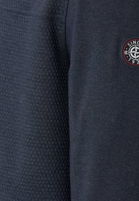 Marineblå sweatshirt med tekstureret, bikube-lignende stof på den ene side og glat materiale på den anden, samt et cirkulært logo badge detalje.
