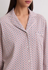 Femme portant une chemise rose à boutons ornée de petits motifs floraux bleus et blancs avec un passepoil blanc autour du col et de la poche.