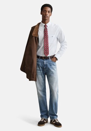 Bruine jas met ribfluwelen kraag, wit-blauw gestreept overhemd, rode stropdas met patroon, lichtblauwe spijkerbroek, bruine sneakers.