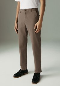 Brown cotton trousers with a straight-leg fit, featuring front pockets, a button closure, and paired with black lace-up shoes.