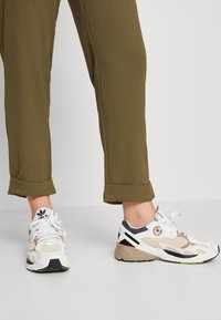 Sportsneakers in wit, beige en zwart met textuurelementen en een contrasterende zool. Draagt bij olijfgroene opgerolde broekspijpen.