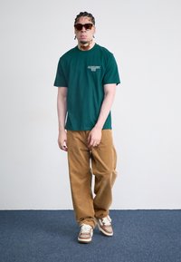 Grønn bomull t-skjorte med grafisk logo, sammen med brune corduroy vide bukser og brune joggesko. Tilbehør inkluderer solbriller.