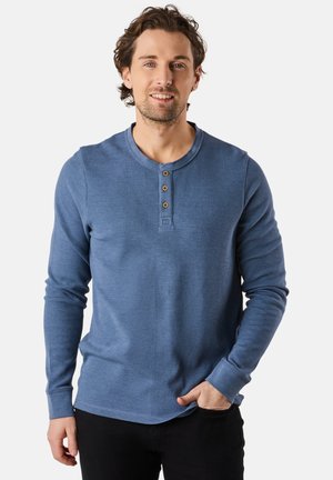 Camicia henley a maniche lunghe blu realizzata in tessuto strutturato, con un colletto rotondo e tre bottoni di legno sul davanti. Indossati pantaloni neri.