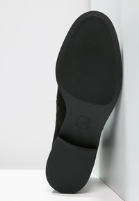 Semelle de chaussure en suede noir avec une texture en caoutchouc antidérapant. Forme arrondie et design plat, avec un logo circulaire embossé au centre.