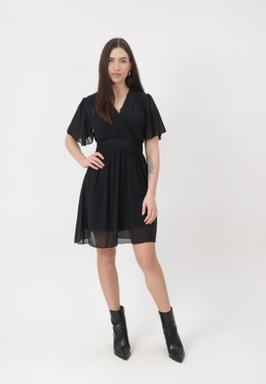 Femme aux longs cheveux foncés portant une robe noire à manches courtes et des bottines noires, debout avec une main sur la hanche sur un fond blanc.