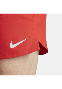Punaiset urheilushortsit, joissa on valkoinen Nike-logo, kulkevat henkilön yläreisillä ja näyttävät kankaan tekstuurin sekä sivuhalkion yksityiskohdan.