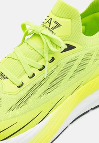 Ljusa neon gula sneakers med stickad överdel, svarta accenter och snörningsdesign. Har texturerade material och en vadderad sula.