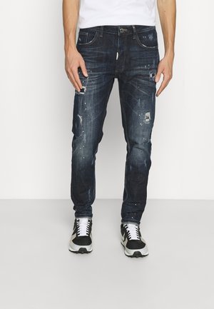 Donkerblauwe spijkerbroek met versleten accenten, waaronder scheuren en verfspatten. Strak model, gecombineerd met zwart-witte sportschoenen.