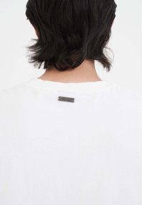 T-shirt in cotone bianco con colletto rotondo e una piccola etichetta nera sopra il colletto posteriore, con una texture liscia.