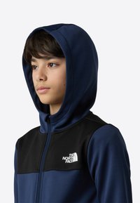 Marineblå hettejakke med glidelås, svarte skuldre og hette, med en liten hvit logo på brystet. Glatt tekstur og vanlig passform.