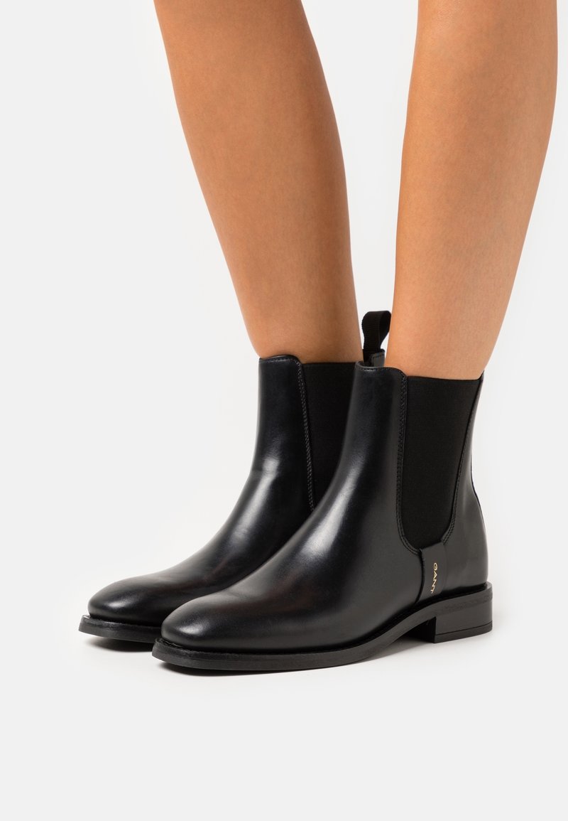 Bottines noires en cuir brillant avec panneaux latéraux élastiques portées sur des jambes nues sur un fond blanc uni.