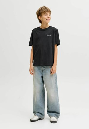 Lachende jongen staand, gekleed in een zwart T-shirt met korte mouwen, lichtblauwe wijde jeans en witte sneakers tegen een effen lichte achtergrond.