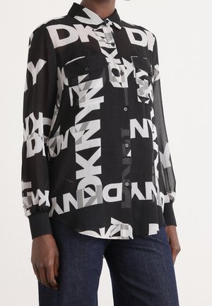 Czarna koszula zapinana na guziki z białymi grubymi literami logo DKNY na całej powierzchni, długie rękawy, kieszenie na piersi, noszona z ciemnoniebieskimi jeansami.