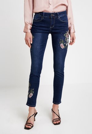 Jeans de denim azul oscuro con corte slim, con bordados florales en las piernas, terminados con sutiles tachuelas para un detalle adicional.