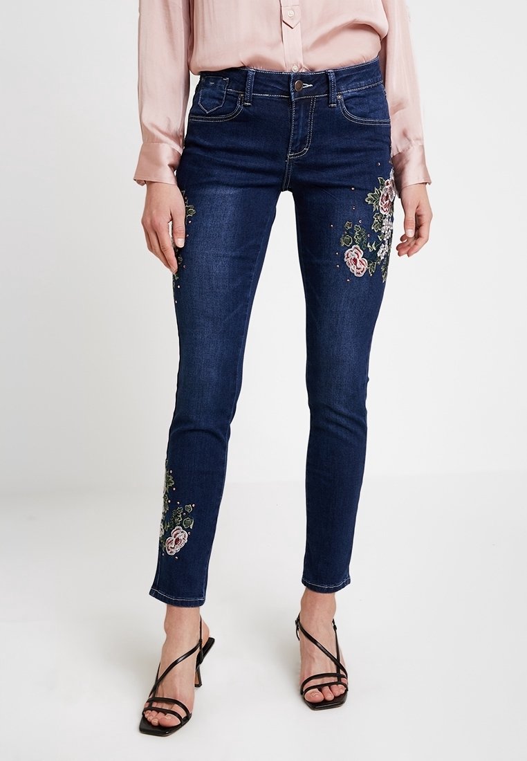 Mörkblå denimjeans med slim fit, prydda med blommig broderi på benen, avslutade med subtila nitar för extra detalj.