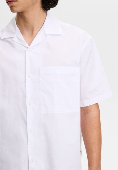 Mężczyzna ubrany w prostą, białą, krótkorękawową koszulę z kołnierzykiem w stylu camp oraz kieszenią na piersi, zwrócony lekko w lewo na jasnoszarym tle.