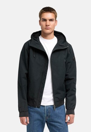 Giovane uomo che indossa una giacca nera con cappuccio e zip sopra una t-shirt bianca e jeans blu su uno sfondo grigio chiaro uniforme.