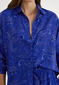 Γυναίκα που φοράει μακρυμάνικο μπλε πουκάμισο με κουμπιά, με λεπτό σχέδιο παϊέτας και μανίκια που έχουν τυλιχτεί.