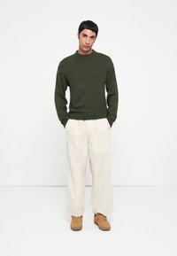 Donkergroene gebreide trui met een geribde halslijn, gecombineerd met losse beige broek en tan sneakers, wat een ontspannen uitstraling creëert.