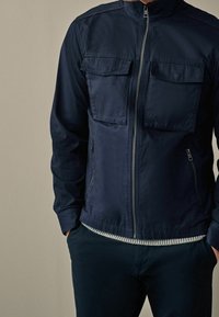 Mężczyzna w granatowej kurtce zapinanej na zamek, z klapkowymi kieszeniami na piersi i bocznymi kieszeniami na zamek, z rękami wsuniętymi w kieszenie spodni na jednolitym beżowym tle.