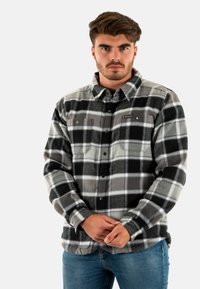 Camicia in flanella a quadri grigia e nera con chiusura a bottoni, due tasche frontali e colletto, indossata sopra dei jeans blu.