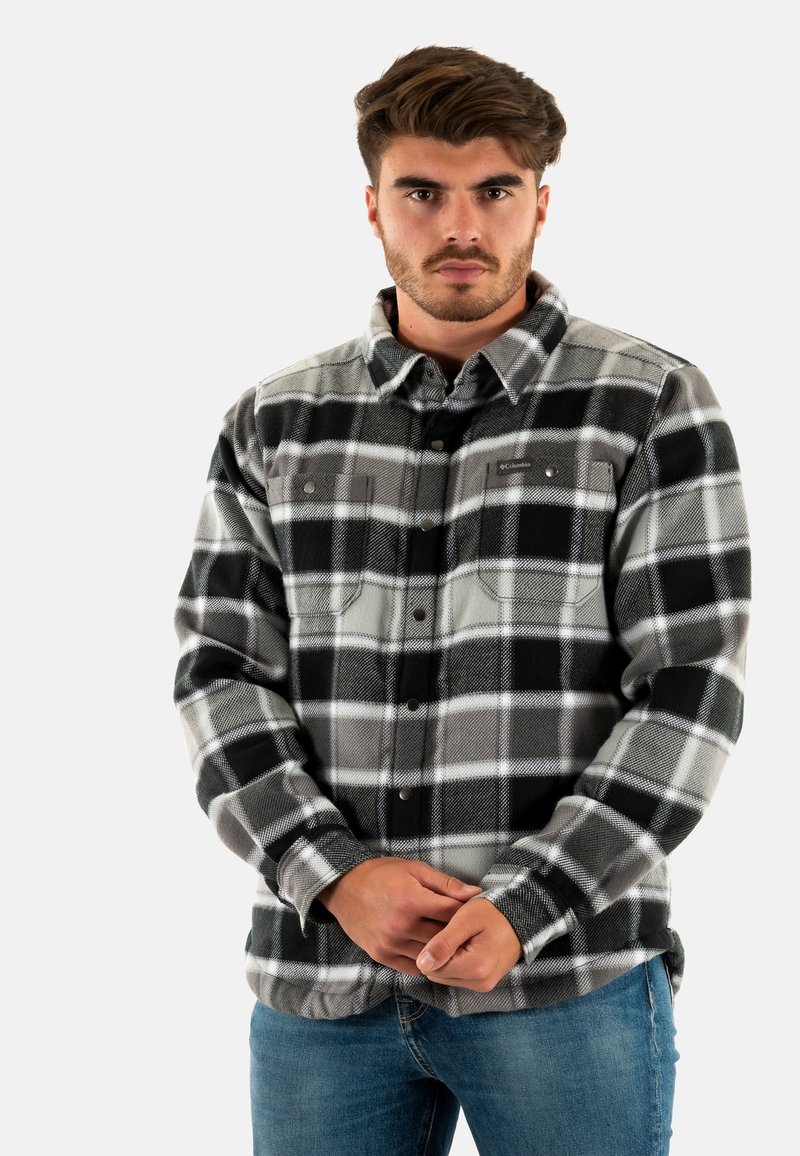 Camicia in flanella a quadri grigia e nera con chiusura a bottoni, due tasche frontali e colletto, indossata sopra dei jeans blu.