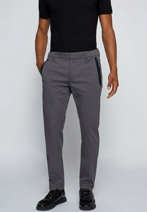 Homme portant un pantalon gris slim avec poches zippées noires, un t-shirt noir et des baskets noires, debout les mains près des poches.