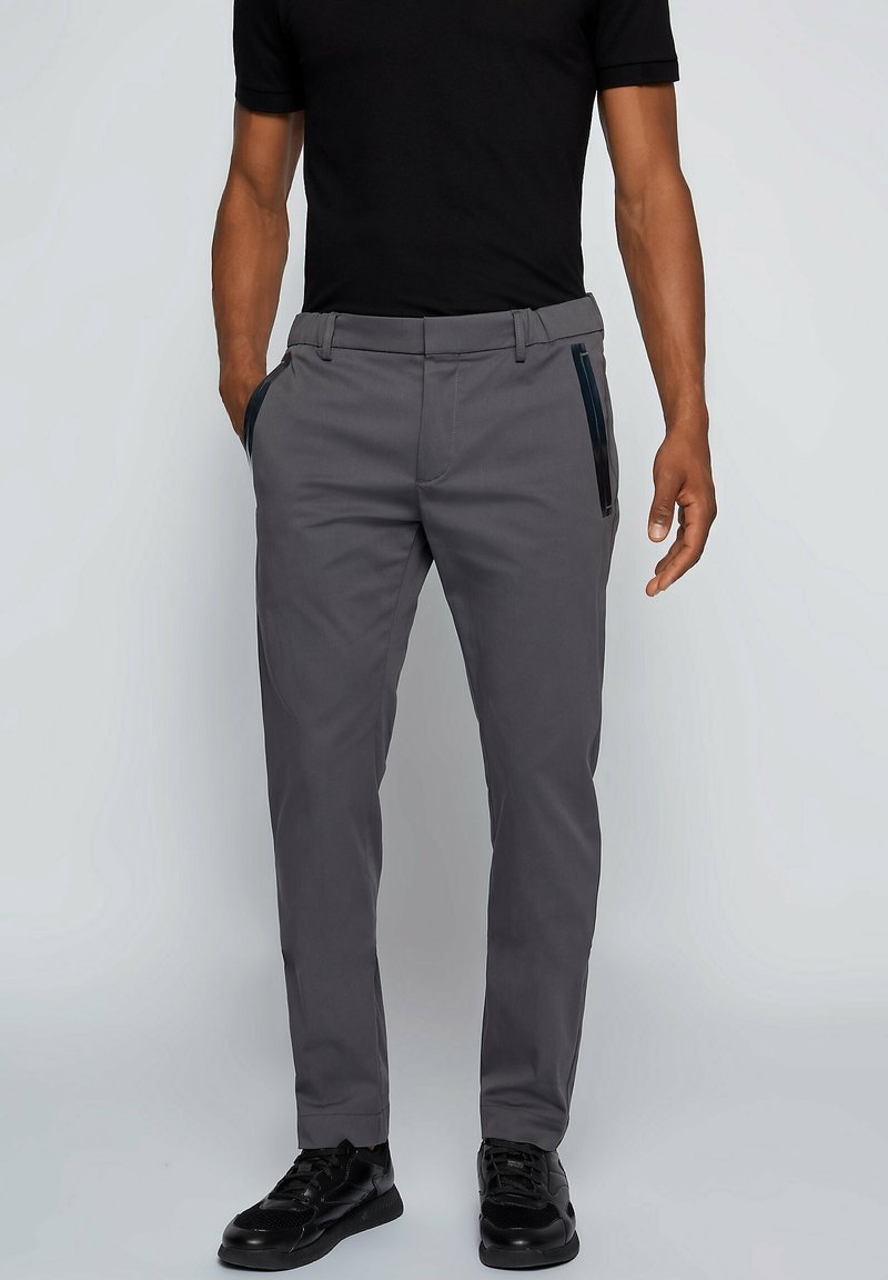 Homme portant un pantalon gris slim avec poches zippées noires, un t-shirt noir et des baskets noires, debout les mains près des poches.